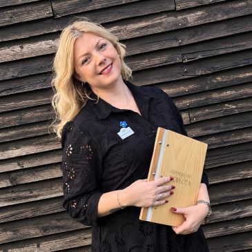 Woman in black dress holding a wooden board, smiling in front of dark wood wall.