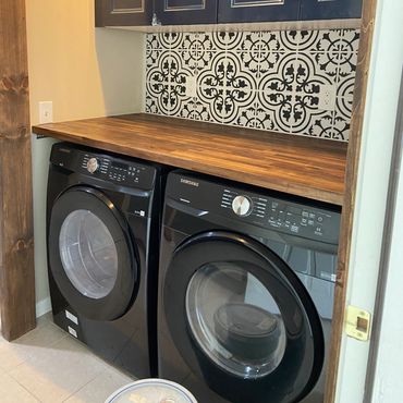 Modern laundry area with black Samsung washer and dryer under wooden countertop and patterned backsplash.
