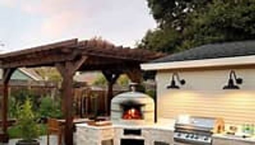 Modern outdoor kitchen with pizza oven, grill, and pergola at sunset.