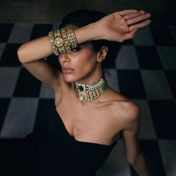 Woman in elegant black dress wearing ornate gold jewelry covering her eyes with arm.