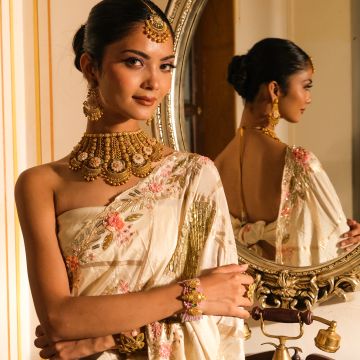 Elegant woman in traditional Indian attire with gold jewelry stands before a mirror.