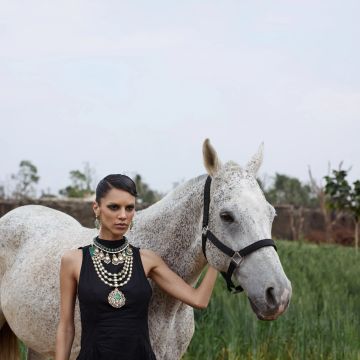 A woman in a black dress with elaborate jewelry stands beside a white horse in a natural setting.