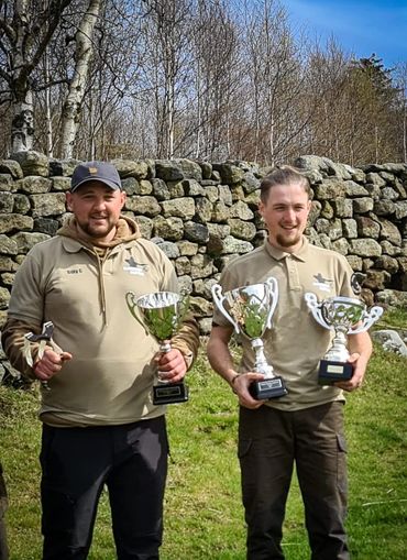 Two men holding trophies outside by a stone wall.