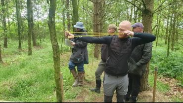 Man using slingshot in forest with observers.