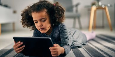 A young girl lying on a striped rug, focused on a tablet.