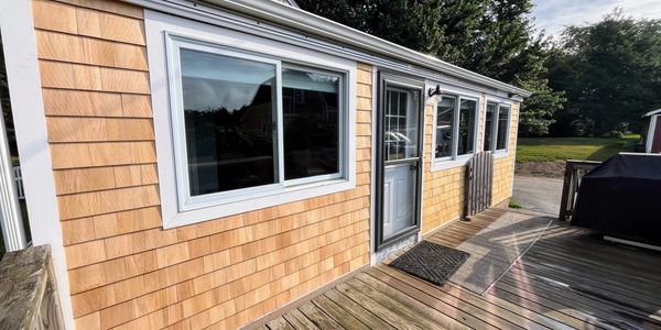 Sunlit wooden deck with a door and windows on a beige shingled house.