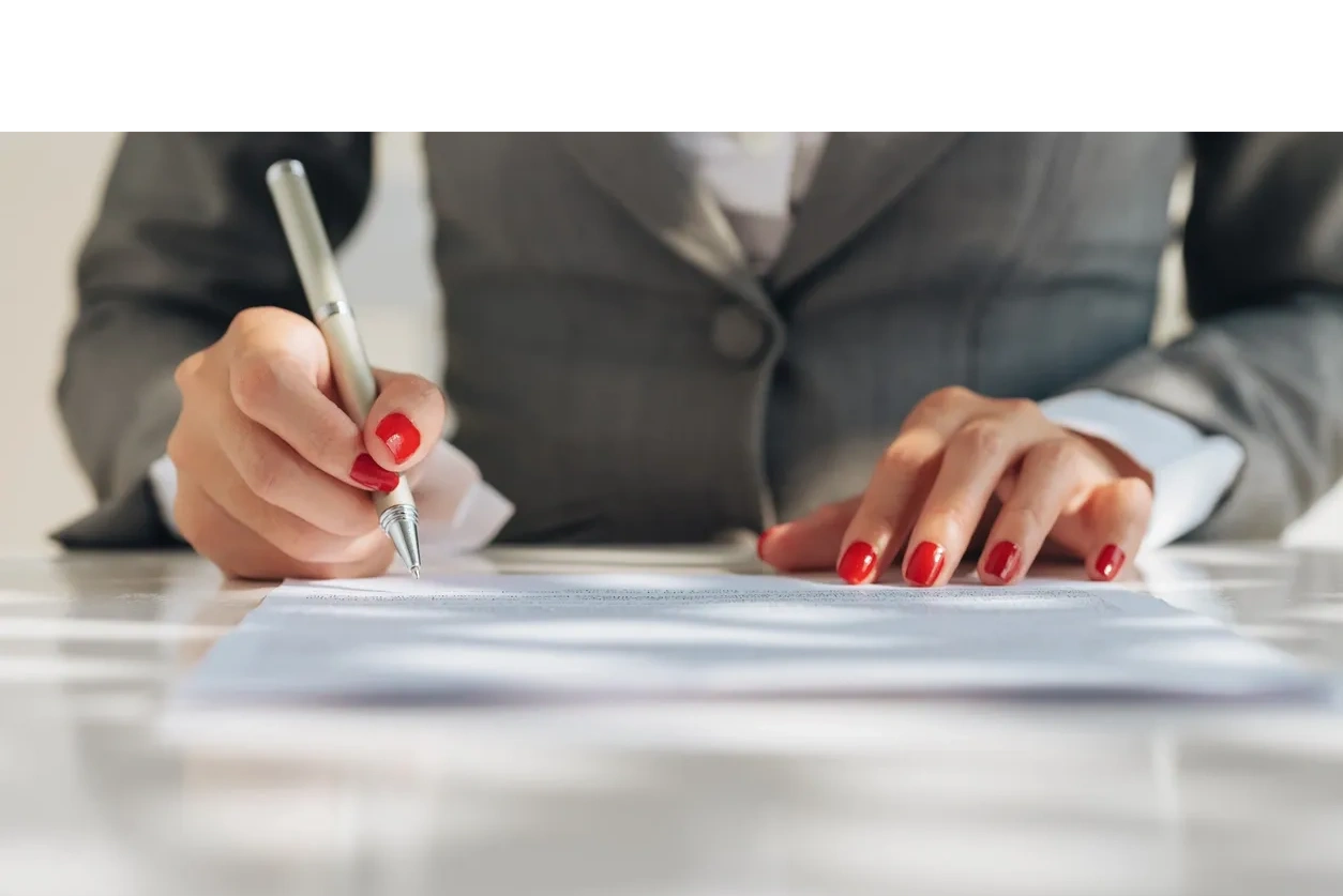 A businesswoman's hands with red nail polish signing an contract with a white pen.