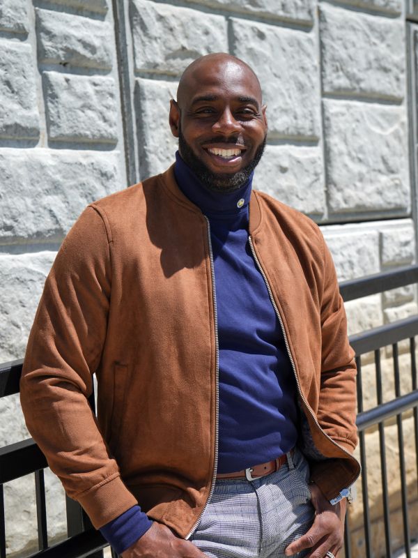 Smiling man in brown jacket and blue turtleneck poses outdoors.