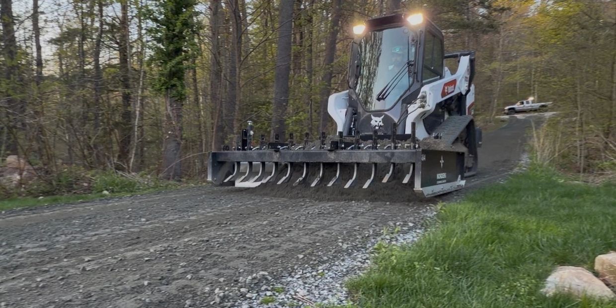 A Bobcat machine grading a dirt and gravel road in a wooded area.