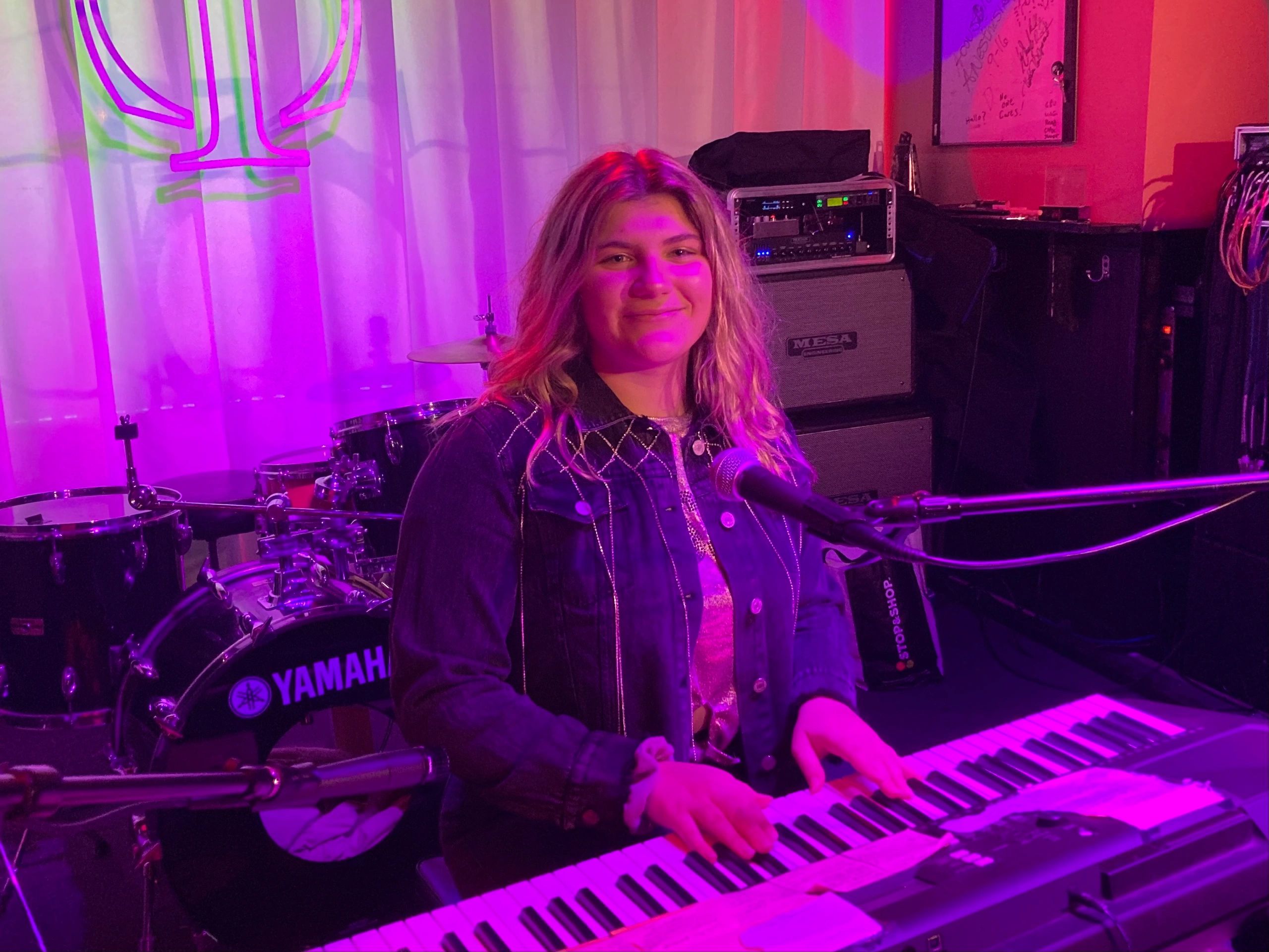 Teenage girl with blonde hair playing keyboard in front of microphone. Yamaha drum set is behind her