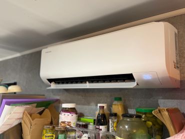 Samsung air conditioner mounted above kitchen shelves with jars and bottles.