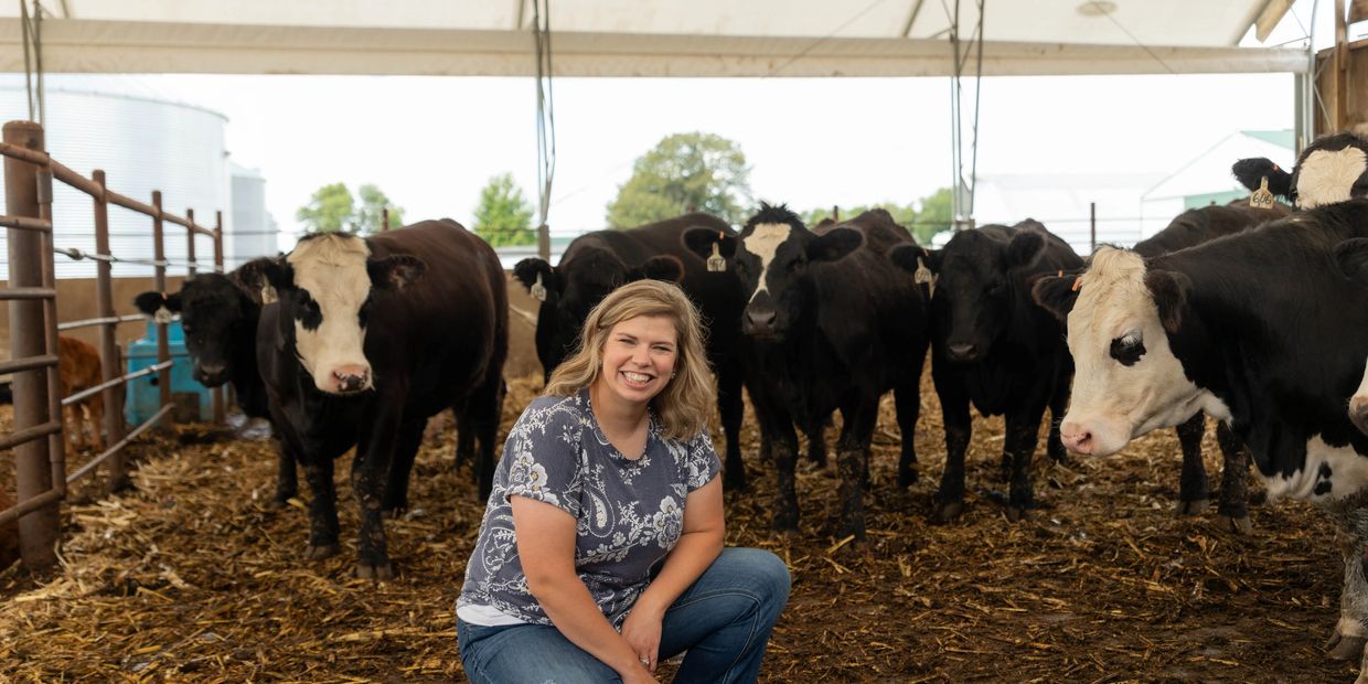 A woman smiling with cows in a barn, under a shelter on a farm.