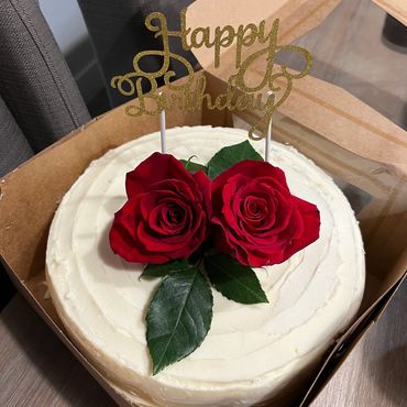 Elegant white birthday cake with two red roses and a golden "Happy Birthday" topper.