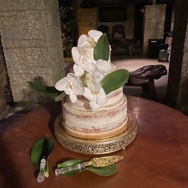 Two-tier semi-naked cake decorated with white orchids and green leaves on a golden stand.