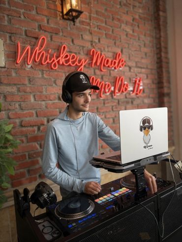 A DJ wearing headphones and a cap is mixing music with a neon sign behind.