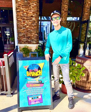 DJ in turquoise shirt and sunglasses points to a colorful brunch event sign.