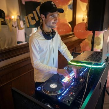 A DJ in a white shirt and cap mixes music at a decorated party venue.