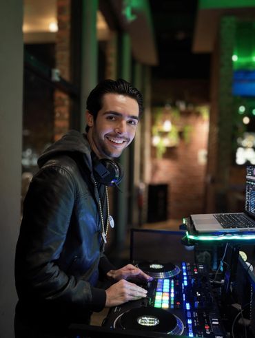 A smiling DJ operating a DJ Controller with colorful lights in a dimly lit room.