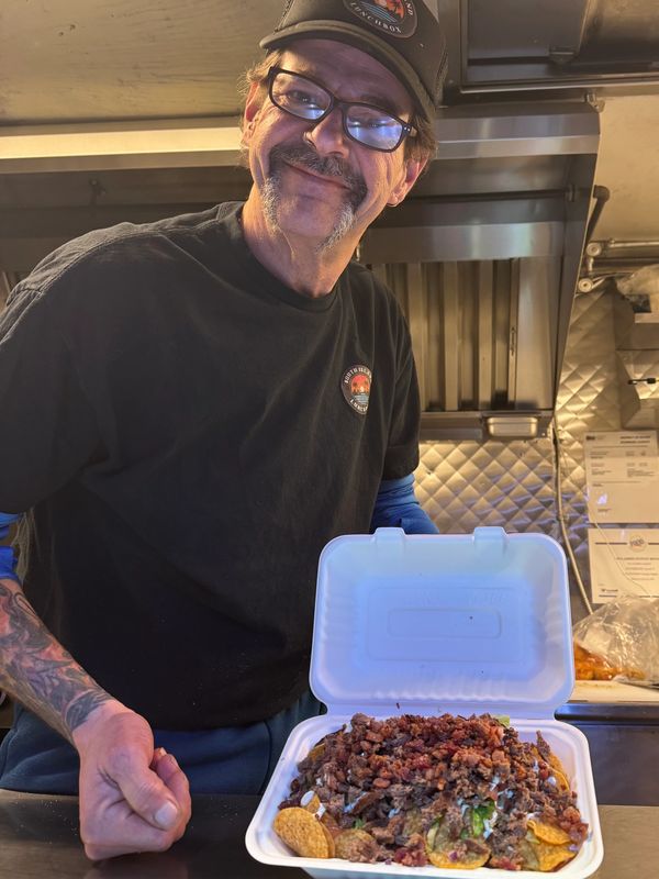 Man smiling and presenting a loaded plate of nachos in a kitchen.