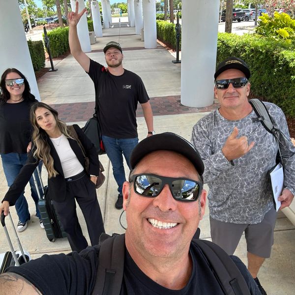 Group of five people smiling outdoors, ready for travel with luggage.