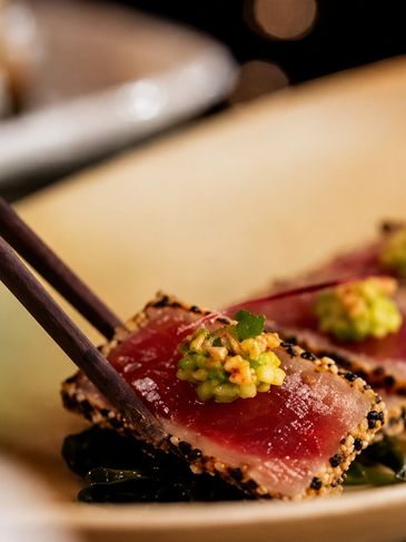 Seared tuna with sesame crust and a dollop of garnish, held by chopsticks.