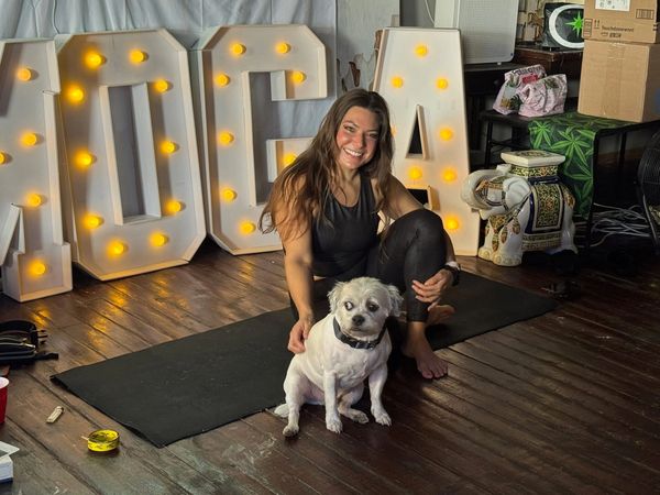 A woman in black workout clothes sits on a mat with her small white dog in a cozy room with lit marquee letters.