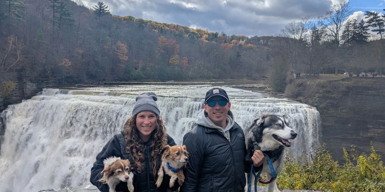 Couple with three dogs posing in front of a large waterfall on a cloudy day.