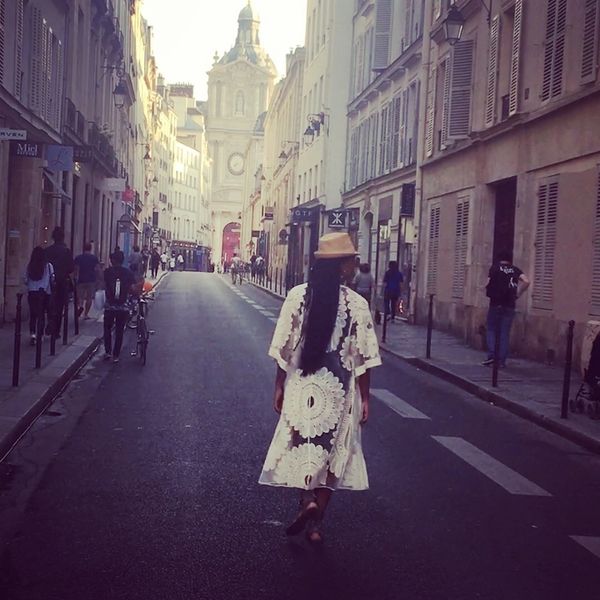 A woman in a white lace dress and hat walks down a historic European street.
