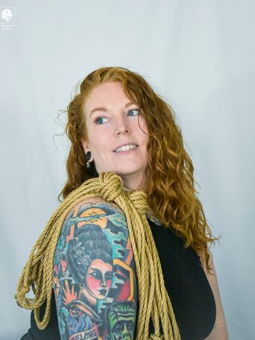 An attractive, red-haired person with tattoos, a black shirt, and rope on their shoulder is smiling.