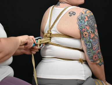 A White, tattooed person with ropes tied on their torso is having an upline & carabiner attached.
