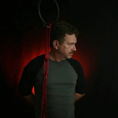 Black & red background; a brunette man wearing a gray shirt stands behind a dangling red rope