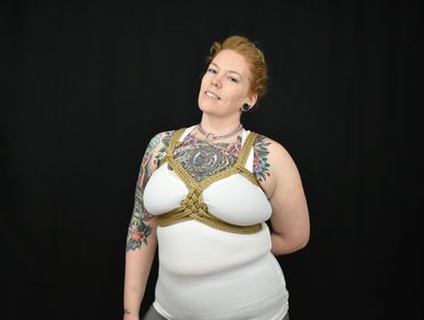 A White, red-headed, fem-presenting person with tattoos on their chest and upper arm, & torso tied.