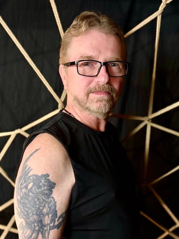 A middle-aged white man with brown hair, a beard & glasses, wearing a black shirt; black background.