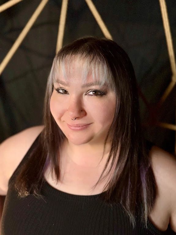 A white woman with white-streaked brown hair wearing a black shirt & nose ring smiles at the camera.