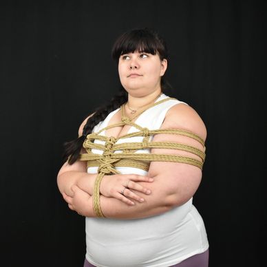 Black background; brunette femme in a white shirt is tied in ropes with their arms crossed.