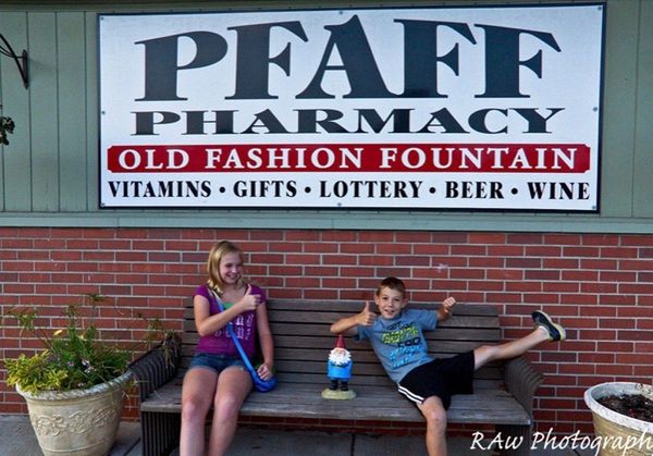 Image: two children sit on bench in front of Pfaff Pharmacy sign giving the thumbs up sign