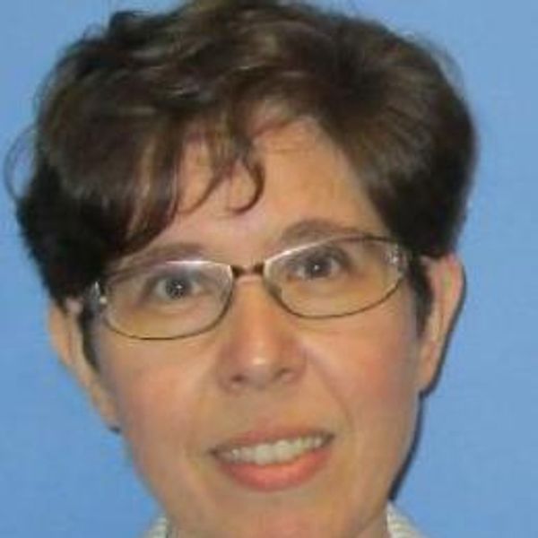 Portrait of a woman with glasses and short brown hair against a blue background.