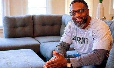 A man wearing an Army shirt smiles, sitting comfortably on a gray couch in a bright living room.