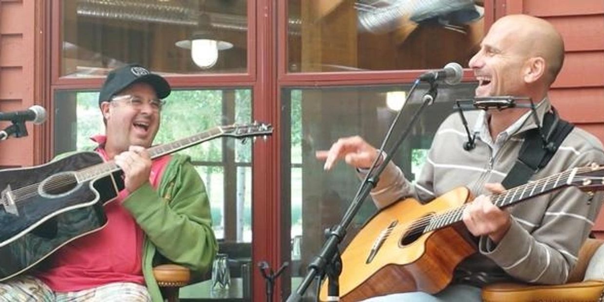 Two men joyfully play guitars and sing together indoors.