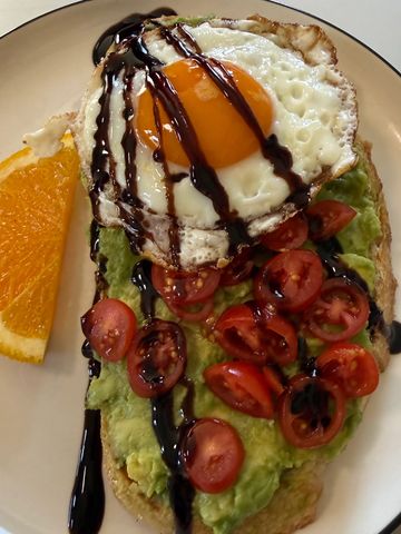 Avocado toast topped with fried egg, cherry tomatoes, and balsamic glaze, served with orange slice.