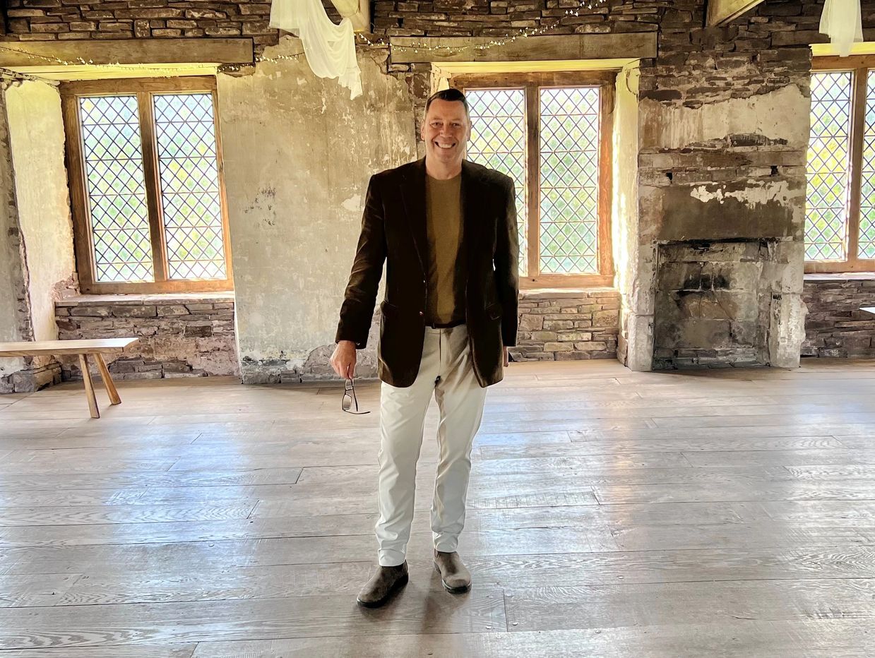Man smiling in a rustic room with old stone walls and wooden floors.