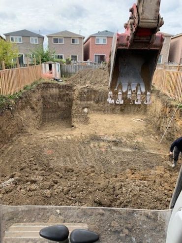 Excavation in a residential backyard with an excavator bucket in view.