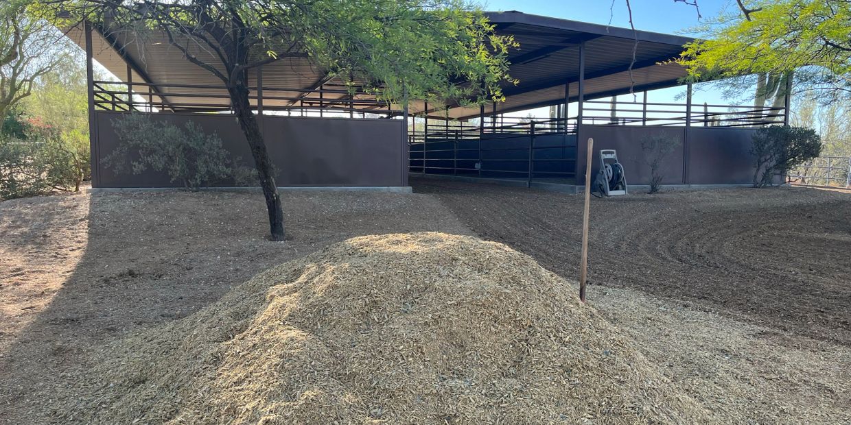 A wood chip pile in front of a covered outdoor stable or animal pen.