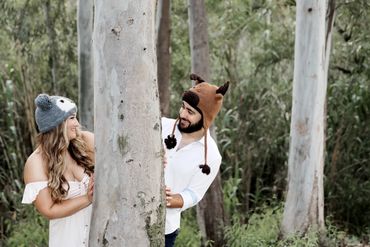 Cute photo of a couple wearing silly knit animal hats peeking around a birch tree at eachother
