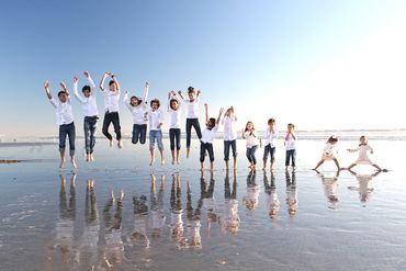 Photo of 14 children in a line in the ocean jumping high in the air. They are reflected in the water