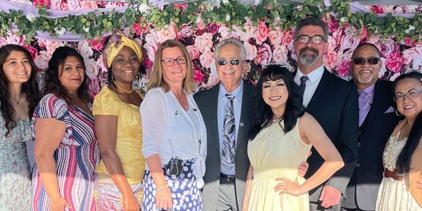 A diverse group of nine people smiling in front of a floral backdrop.