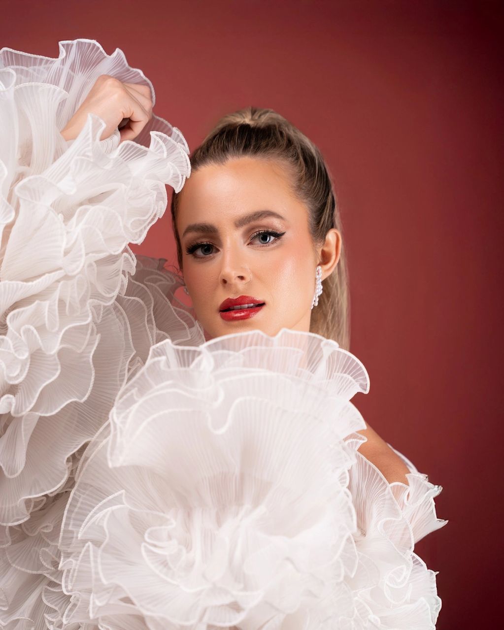 Woman in a ruffled white outfit poses against a red background.