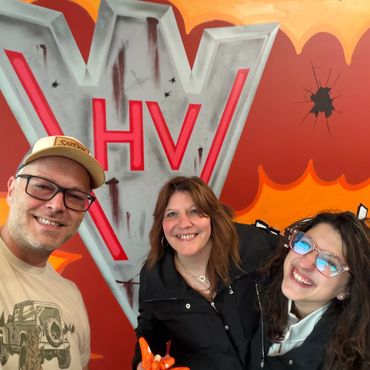 Three smiling people posing in front of a colorful graffiti wall with the letters 'HV'.