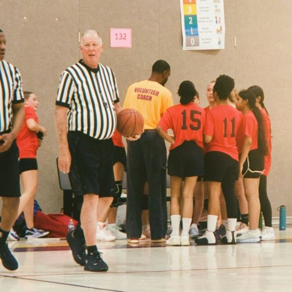 Referee holding basketball near a girls' volleyball team and volunteer coach.