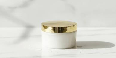 White cosmetic jar with a gold lid on a marble surface.